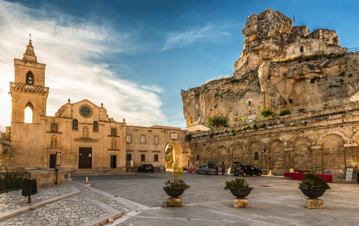 Platz vor einer historischen Kirche und Felsen mit blauen Himmel im Hintergrund.