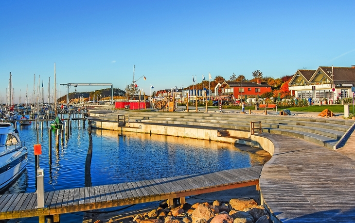 Hafen mit Segelbooten, Steg und Gebäuden am Ufer unter klarem blauem Himmel bei sonnigem Wetter.