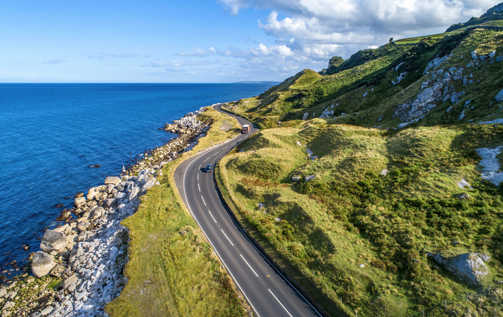 Küstenstraße mit Autos neben grünen Hügeln und blauem Meer bei klarem Himmel.