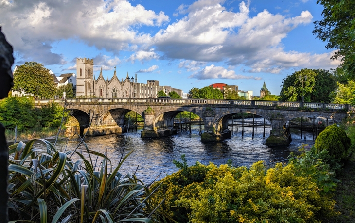 Steinbrücke über einen Fluss mit Kirche im Hintergrund und grüner Vegetation im Vordergrund