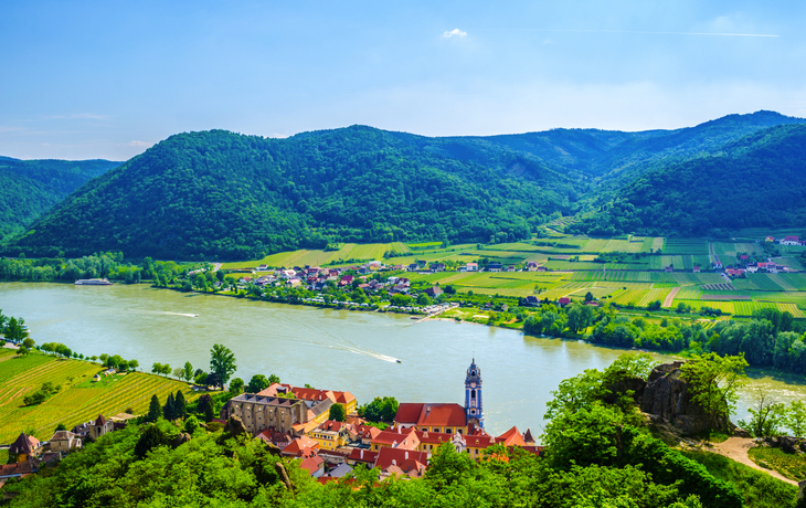 Landschaftsansicht eines Flusses, umgeben von grünen Hügeln und einem kleinen Dorf mit einer markanten Kirche im Vordergrund.