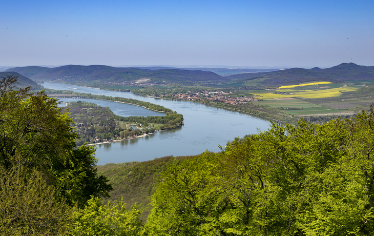 Landschaft mit Fluss und bewaldeten Hügeln unter blauem Himmel.