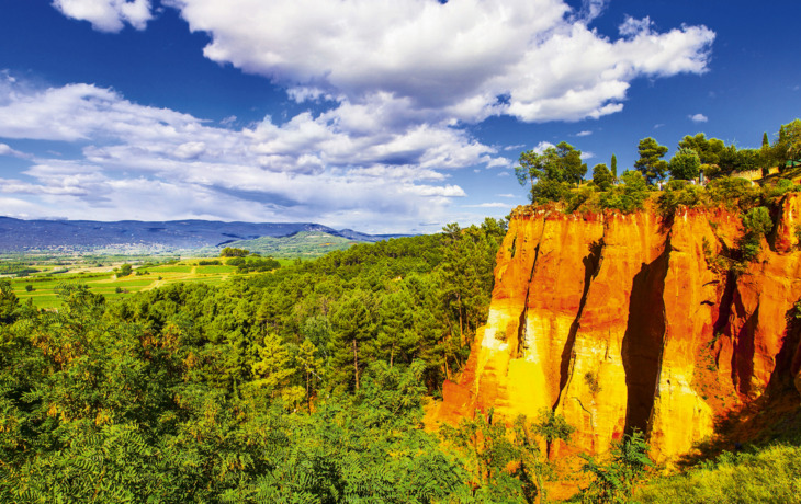 Bunte Sandsteinfelsen vor bewaldeter Landschaft und blauem Himmel mit Wolken.