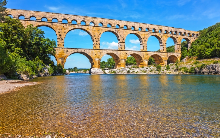Römisches Aquädukt mit mehreren Bögen über einem Fluss an einem sonnigen Tag mit blauem Himmel.