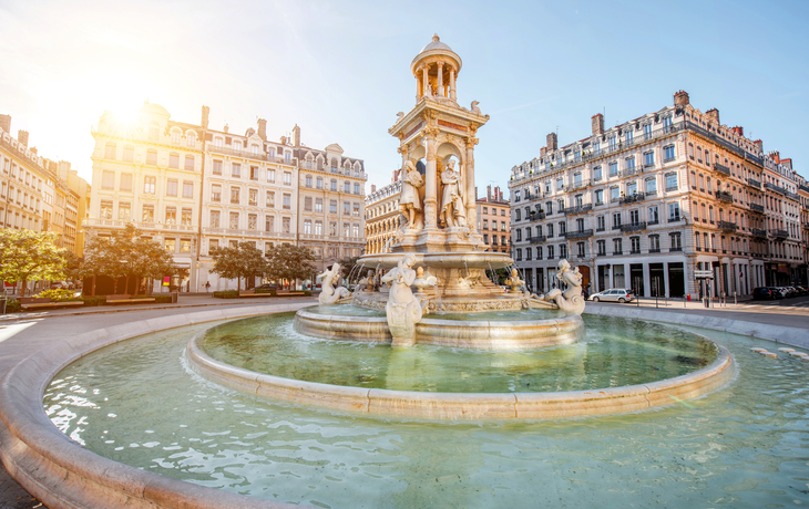 Brunnen auf einem Platz mit historischen Gebäuden im Hintergrund bei Sonnenschein.