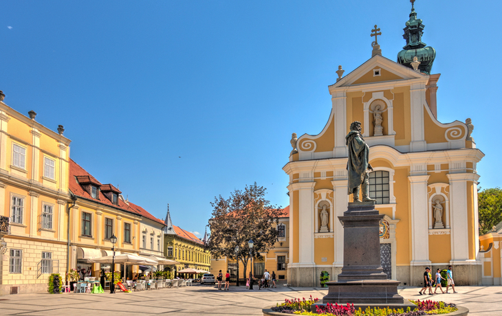Platz mit Statue, barocker Kirche und Gebäuden unter blauem Himmel.