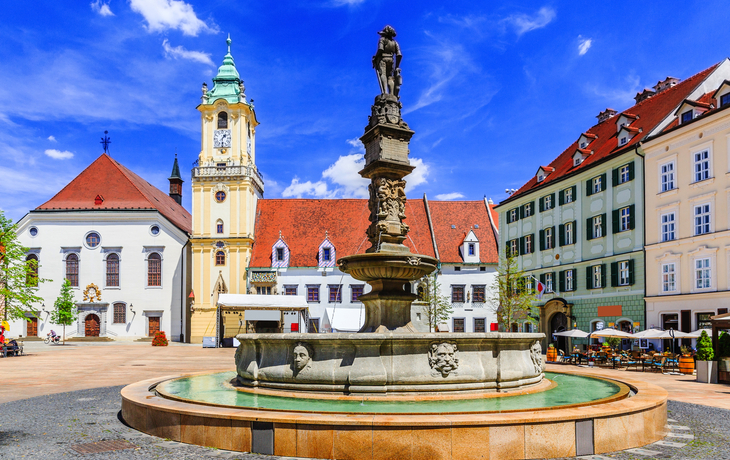 Historischer Platz mit Brunnen und Gebäuden unter klarem Himmel.
