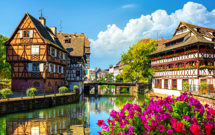 Fachwerkhäuser an einem Kanal in Straßburg bei sonnigem Wetter mit Blumen im Vordergrund.