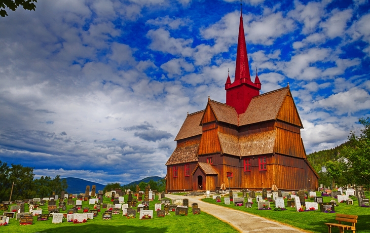 Holzkirche mit rotem Turm und Grasdach, umgeben von einem Friedhof, unter blauem Himmel mit Wolken.