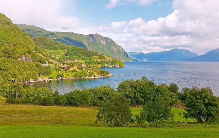 Landschaft mit Fjord, Bergen und Wiesen bei bewölktem Himmel.