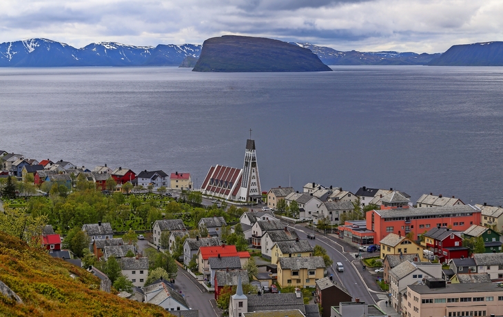 Küstenstadt mit Kirche, Bergen und Insel im Hintergrund unter bewölktem Himmel.