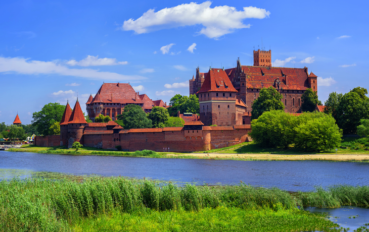 Burg auf Hügel am Flussufer unter blauem Himmel mit Wolken.