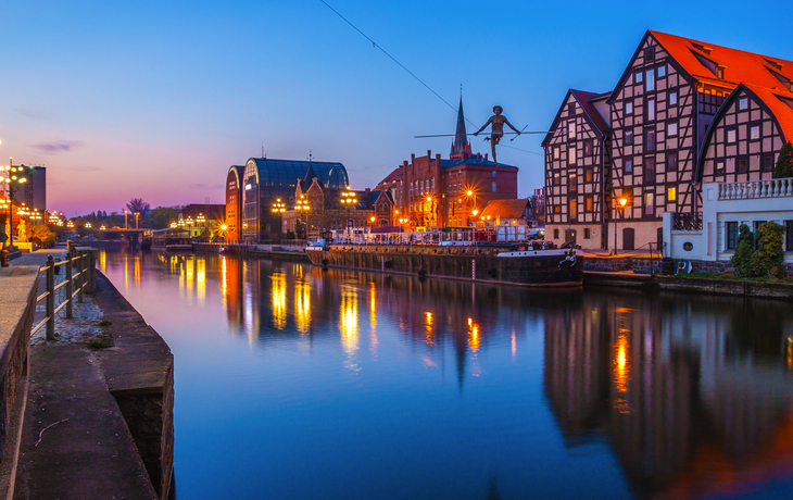 Stadtansicht bei Dämmerung mit Fachwerkhäusern an einem Flussufer.