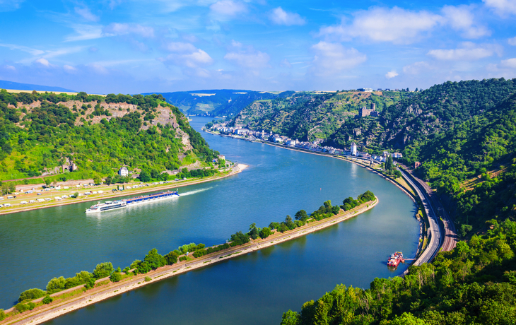 Flusslandschaft mit Hügeln, Bahngleisen und Booten bei sonnigem Wetter