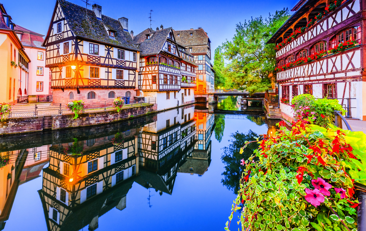 Fachwerkhäuser am Wasser mit Blumen im Vordergrund und spiegelnder Wasserfläche.