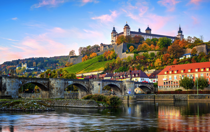 Festung auf einem Hügel mit Brücke im Vordergrund, herbstliche Bäume im Hintergrund.