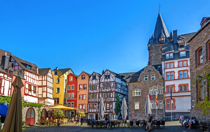 Buntem Fachwerkhäusern umgebener Platz mit Tischen und Sonnenschirmen bei blauem Himmel.