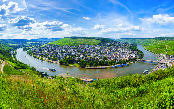 Panoramablick auf eine Flussbiegung mit Stadt und grünen Hügeln unter blauem Himmel.