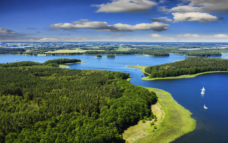 Landschaft mit Seen, grünen Wäldern und weißen Segelbooten unter blauem Himmel.