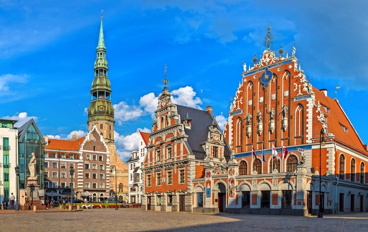 Historisches Gebäude und Kirchturm auf einem Platz bei blauem Himmel.