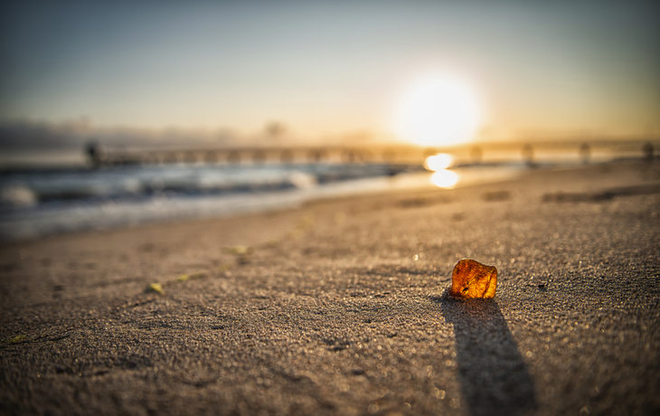 Ein Stück Bernstein liegt im Sonnenaufgang am Strand, im Hintergrund ist das Meer sichtbar.