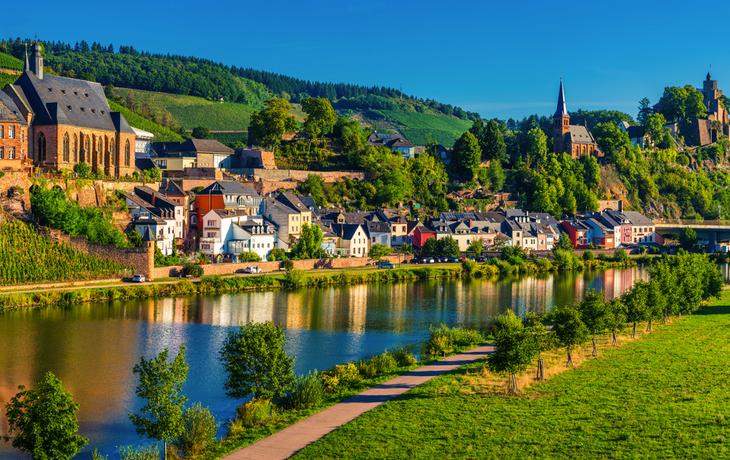 Panorama einer Flusslandschaft mit Dorf und bewaldeten Hügeln im Hintergrund.