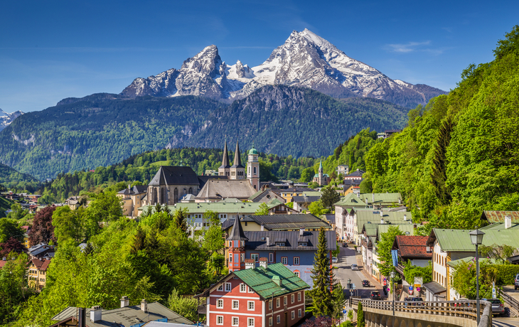 Stadt mit Kirche und Alpen im Hintergrund bei klarem Himmel.