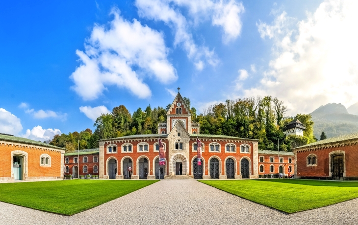Historisches Gebäude mit Bögen und Innenhof vor bewaldetem Hügel.