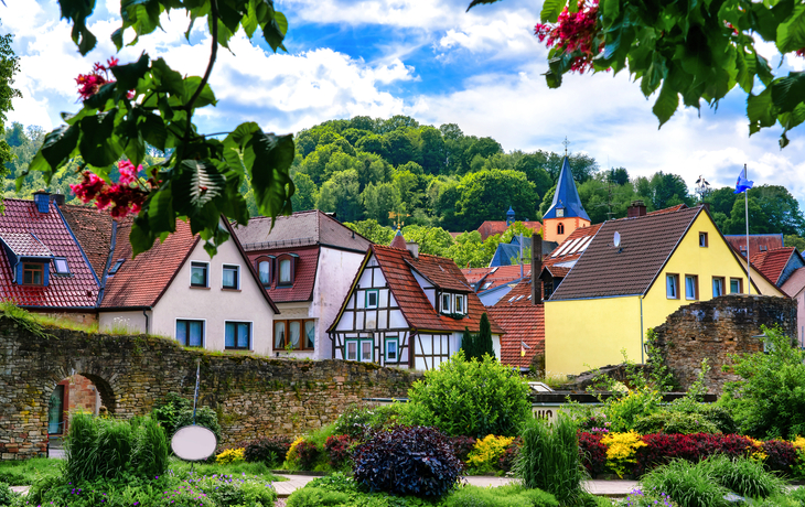 Fachwerkhäuser in einem Dorf mit blühendem Garten und bewaldetem Hügel im Hintergrund.