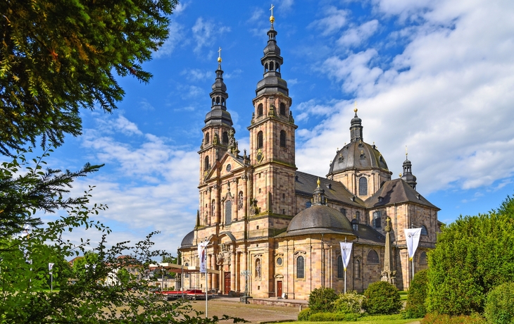 Barocke Kathedrale mit Türmen vor blauem Himmel und umgeben von Bäumen.