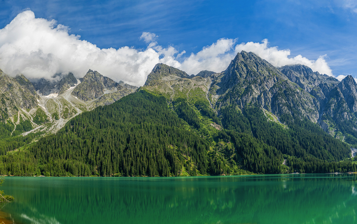 Bergsee mit grüner Wasserfläche und bewaldeten Bergen im Hintergrund bei klarem Himmel.