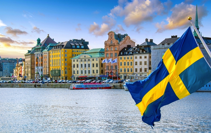 Schwedische Flagge weht vor bunten Gebäuden am Wasser.