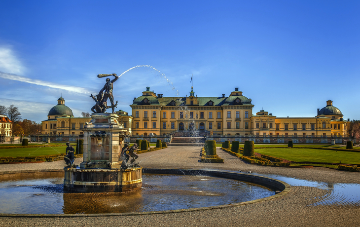 Schloss mit Brunnen im Vordergrund unter blauem Himmel.