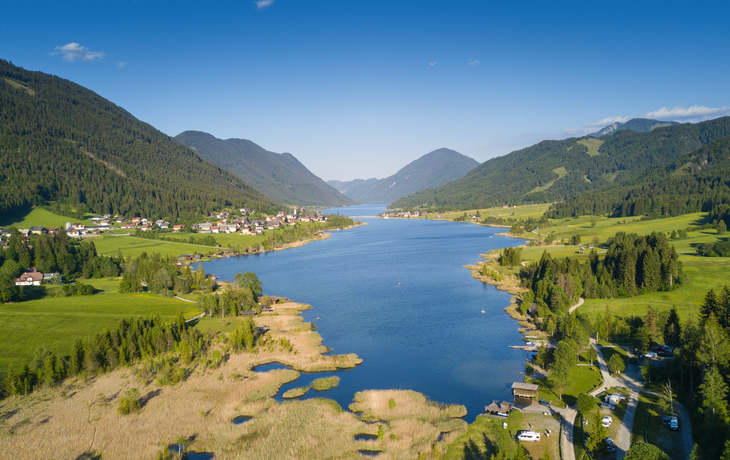 Landschaft mit Bergsee, umgeben von Wiesen und Wäldern unter klarem Himmel.