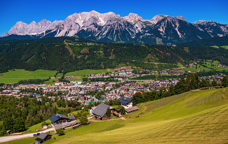 Landschaft mit Bergen, Dorf im Tal, grüne Wiesen, blauer Himmel.