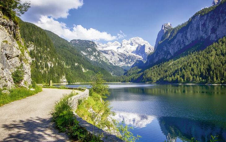 Bergsee mit Wanderweg und schneebedeckten Gipfeln im Hintergrund.