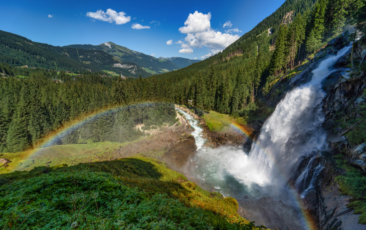 Berglandschaft mit Wasserfall und Regenbogen unter blauem Himmel.