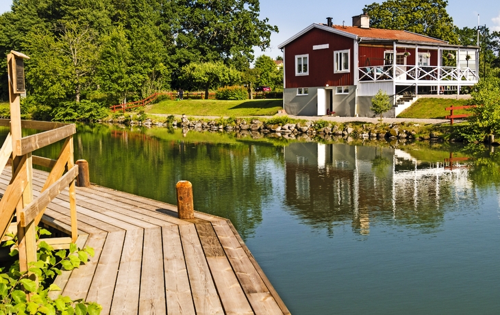 Holzdeck am See mit rotem Haus im Hintergrund und üppiger Vegetation.