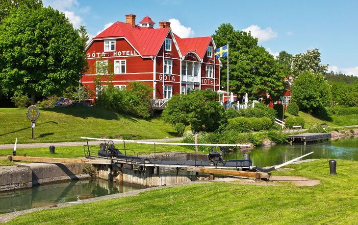 Rotes Hotel mit rotem Dach am Kanal, umgeben von Bäumen und Wiese, mit Schleuse im Vordergrund.