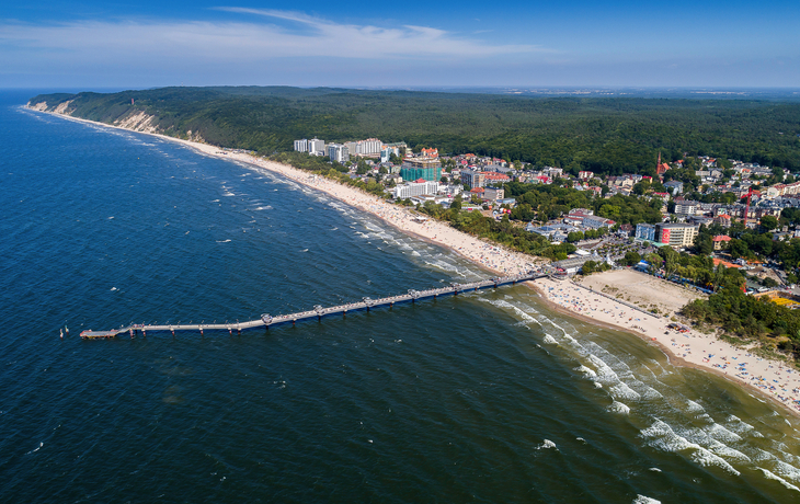 Küstenstadt mit Strand, Pier und grünem Wald aus der Luftansicht.