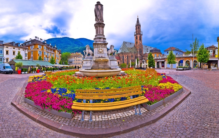 Stadtplatz mit Blumenbeeten, Statue und Kirche im Hintergrund.