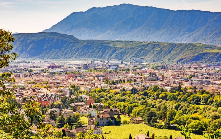 Stadtansicht mit grünen Bäumen und Bergen im Hintergrund bei sonnigem Wetter.