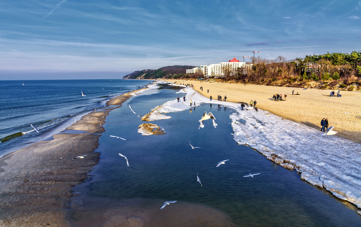 Küste mit Strand, Möwen, Menschen und Gebäuden im Hintergrund.