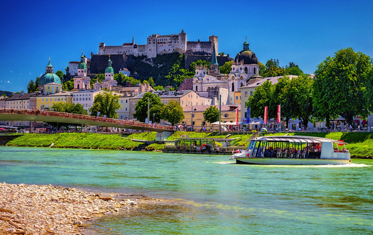 Fluss mit Boot, historische Gebäude und Burg in einer Stadtlandschaft bei klarem Himmel.