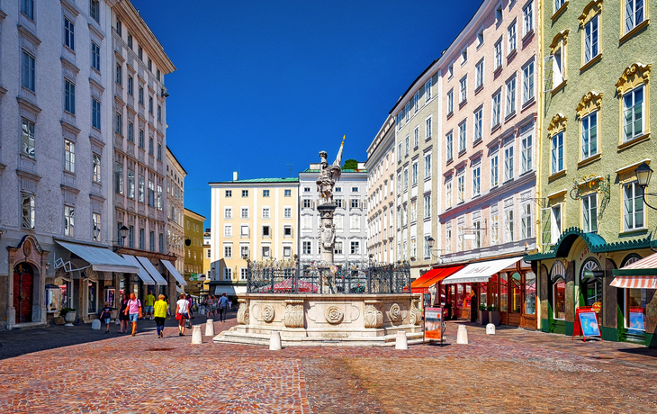 Städtischer Platz mit Springbrunnen und umliegenden Pastellgebäuden bei klarem Himmel.