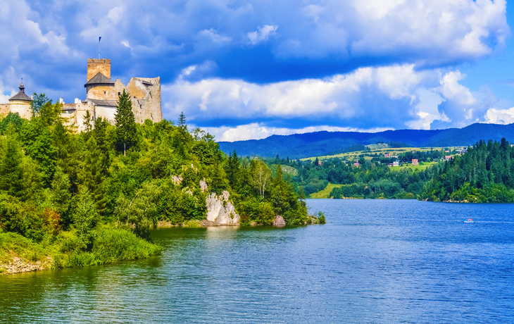 Burg am Seeufer mit bewaldeten Hügeln im Hintergrund unter blauem Himmel.