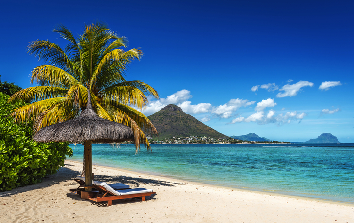 Strand mit Palme, Liege und Sonnenschirm vor tropischer Meereskulisse.