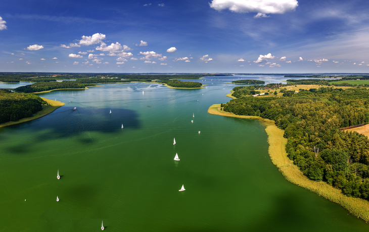 Landschaft mit grünem See, Segelbooten und bewaldeten Ufern unter blauem Himmel