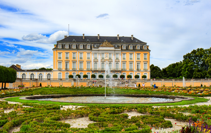 Barockschloss mit gepflegtem Garten und Springbrunnen im Vordergrund.