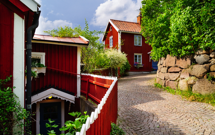 Rote Holzhäuser an einer Kopfsteinpflasterstraße, umgeben von üppigem Grün.